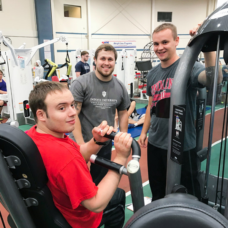 An adult AMP client smiles with two instructors in the gym's strength machine area.