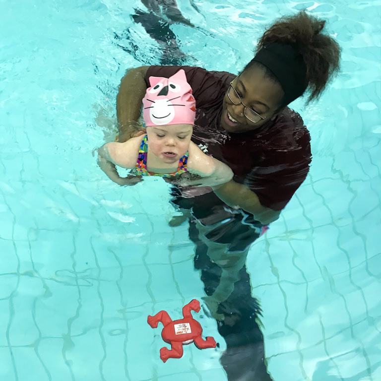 A female instructor helps a toddler in a bathing suit and pink plastic swim cap swim towards a floating toy. 