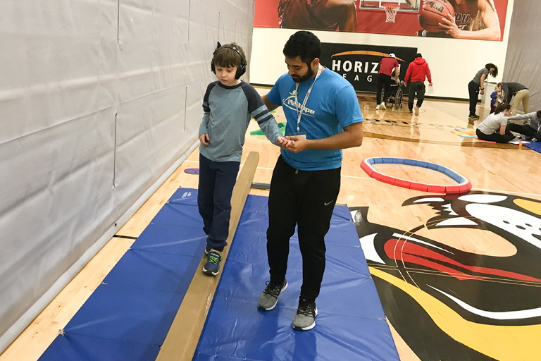 A male AMP instructor helps a boy wearing sound deadening headphones walk along a balance beam in the IUPUI gym.