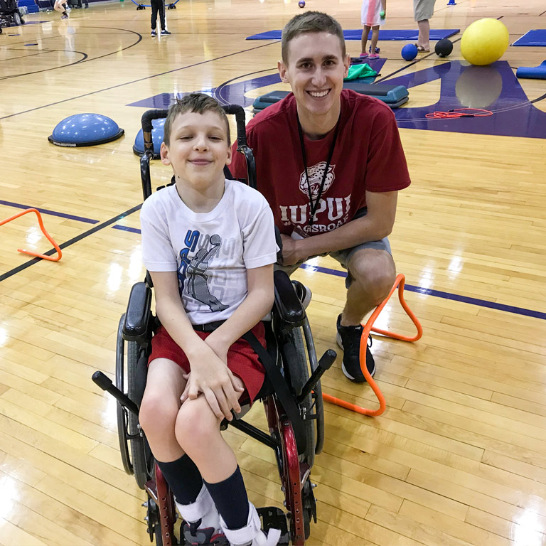 A male instructor in an IUPUI t-shirt poses for a photo with a young AMP client in a sport wheelchair.