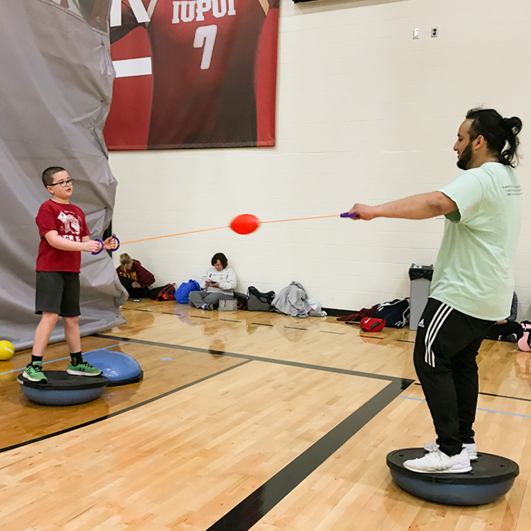 A male instructor and an elementary-aged boy balance together on medicine balls. 