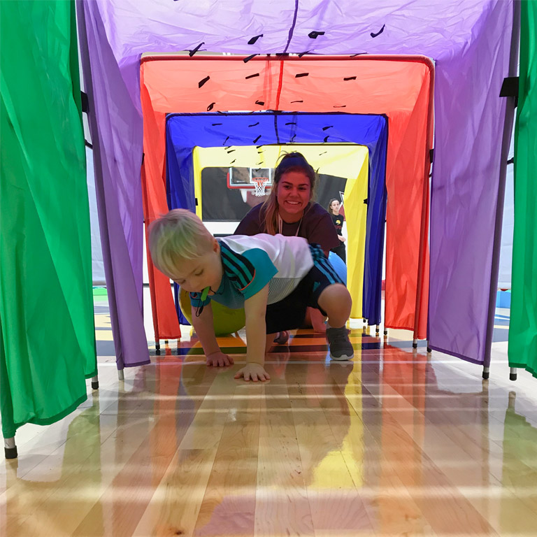 A little boy crawls through a brightly colored fabric tunnel under instructor supervision.